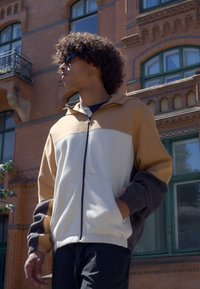 Chaqueta en bloques de color en tonos beige, crema y gris oscuro. Cuenta con un cuello alto, cierre de cremallera y puños elásticos.