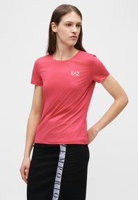 Γυναίκα που φοράει εφαρμοστό ροζ μπλουζάκι με κοντά μανίκια και το λογότυπο EA7, καθώς και μαύρη φούστα με κάθετη λωρίδα κειμένου Emporio, στέκεται με το χέρι στη μέση.