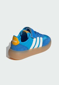 Blue suede sneaker with white stripes and a yellow accent on the heel. Features a rounded toe and a semi-transparent rubber sole.