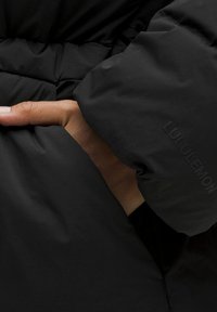 Giacca a vento nera con finitura texturizzata, con una mano in una tasca laterale e un marchio sottile visibile sulla manica.
