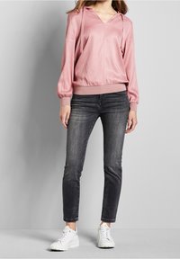 Rosa Satin-Hoodie mit V-Ausschnitt, elastischen Bündchen und Saum. Kombiniert mit dunkelgrauen, eng geschnittenen Jeans und weißen Sneakers.