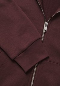 Felpa con zip color bordeaux realizzata in tessuto morbido, dotata di una cerniera metallica e polsini a coste. La texture è liscia con una leggera elasticità.