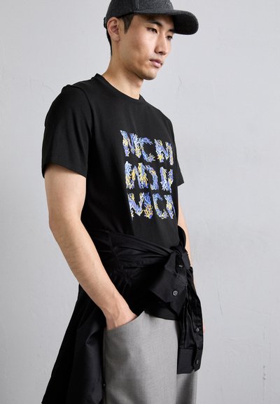 T-shirt noir à manches courtes avec un texte en forme de corail multicolore. Le modèle porte un pantalon gris clair et une chemise noire nouée autour de la taille.
