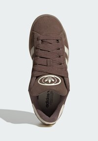 Brown suede sneaker with textured laces, white stripes, and a round logo. Features a cushioned tongue and padded collar for comfort.