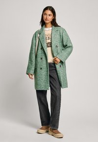 Manteau en laine vert à motif chevrons, coupe oversize, design double boutonnage et grands boutons, porté sur un pull crème et un jean foncé.