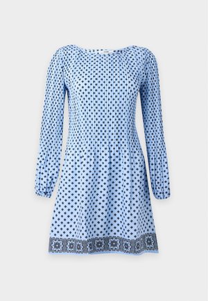 Donna che indossa un vestito a maniche lunghe azzurro chiaro con motivi a diamante blu scuro e orlo floreale, abbinato a calzini bianchi e scarpe marroni.