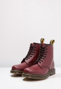 Bottes chevilles en cuir bordeaux avec lacets noirs, surpiqûres jaunes et languettes de tirage noires et jaunes distinctives. Semelle en caoutchouc transparente avec relief antidérapant.