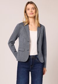 Femme portant un blazer gris cintré sur un haut blanc et un jean bleu foncé, debout contre un fond clair uni, mains détendues.
