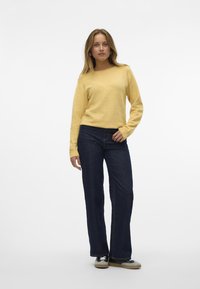 Gul strikket sweater med lange ærmer, parret med mørkeblå flared jeans og sort-hvide sneakers. Glat tekstur, afslappet pasform.