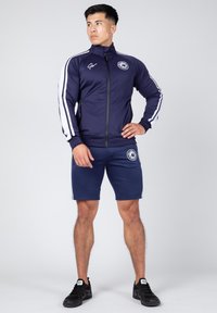 Marineblauwe sportjack met witte strepen en logo, met ritssluiting, gecombineerd met bijpassende shorts. De stof lijkt glad en rekbaar. Zwarte sneakers.