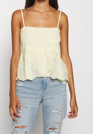 Kvinde iført en bleg gul, broderet top med spaghetti-stropper kombineret med lyseblå revnede jeans mod en ensfarvet baggrund.