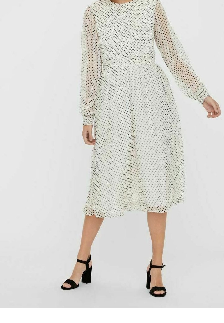Bílé šaty s puntíky, průsvitnými dlouhými rukávy, zvýrazněným pasem a rozšířenou sukní. Délka ke kolenům, zakončeno volánkovým okrajem. Černé lodičky.