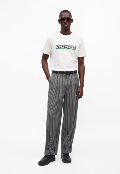 Homme portant des lunettes de soleil, un t-shirt blanc avec un texte graphique, un pantalon gris à fines rayures, une ceinture noire et des chaussures noires, debout avec une main dans la poche.