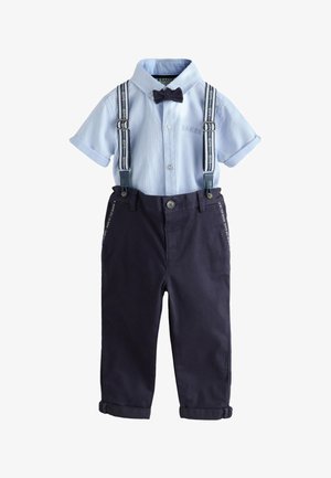Jeune garçon souriant, portant une chemise bleu clair avec un nœud papillon bleu marine, des bretelles bleu marine, un pantalon bleu marine et des baskets blanches, debout sur un sol gris.