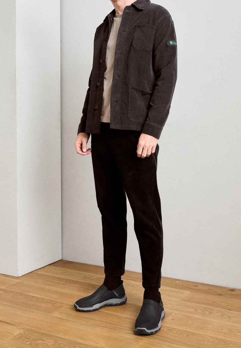 Veste en velours côtelé foncé avec poches avant, associée à un pantalon noir côtelé et des chaussures grises à enfiler, sur un fond uni avec un sol en bois.