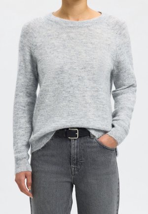 Personne portant un pull gris clair à manches longues, rentré dans un jean taille haute gris foncé avec une ceinture noire, une main dans la poche.