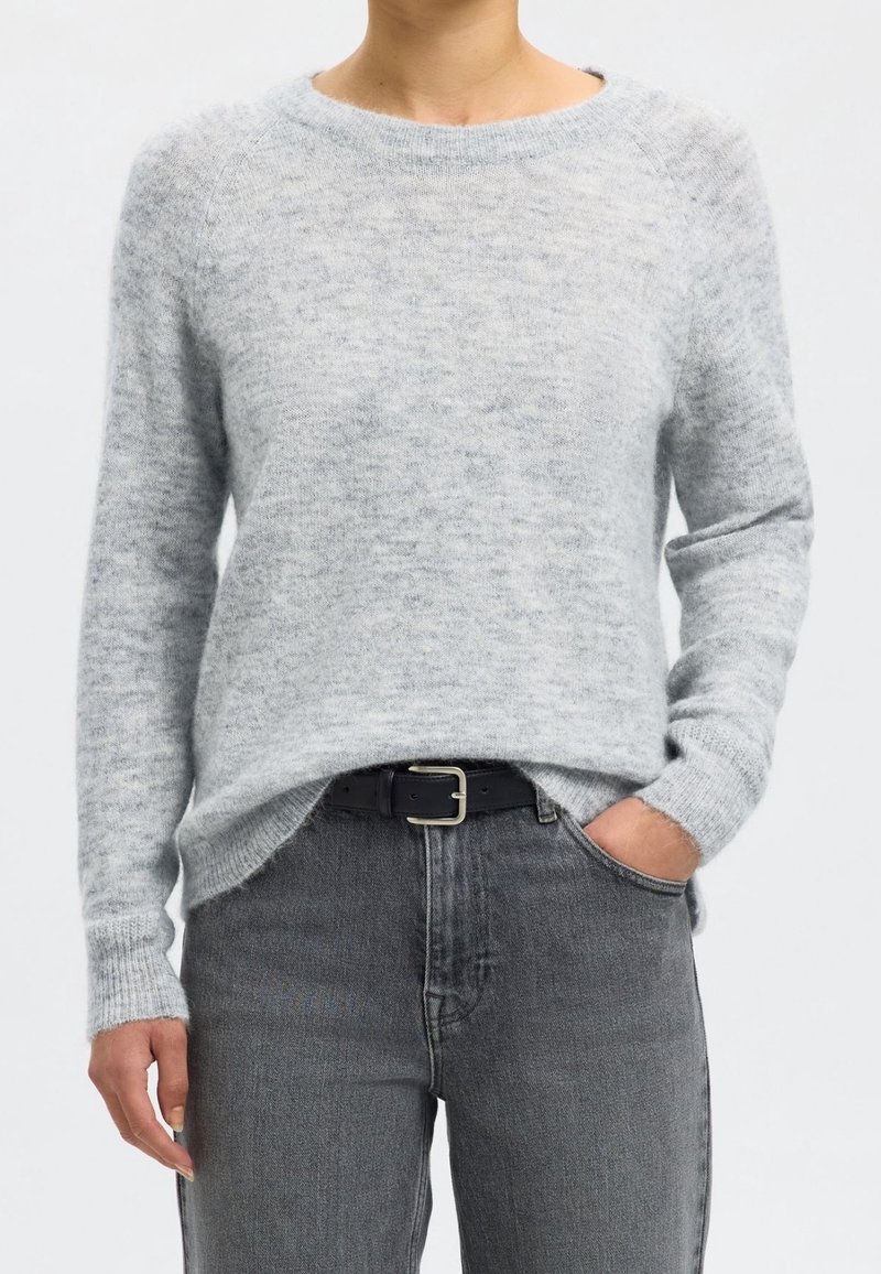 Personne portant un pull gris clair à manches longues, rentré dans un jean taille haute gris foncé avec une ceinture noire, une main dans la poche.