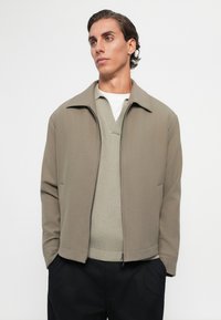 Jeune homme avec des cheveux coiffés en arrière, portant une veste beige zippée sur un pull en mailles léger et un pantalon noir, debout avec les mains dans les poches.