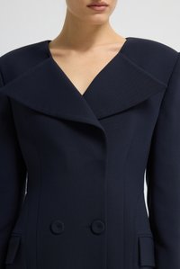 Giacca tailored blu navy con grande revers affilato, design doppiopetto e tessuto testurizzato. Presenta bottoni rotondi e spalle strutturate.