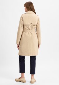 Vrouw in een beige trenchcoat met riem, marineblauwe broek en beige loafers, staand met haar rug naar de camera tegen een effen witte achtergrond.