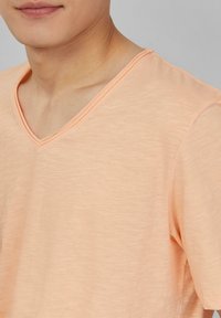 Brzoskwiniowa koszulka typu V-neck wykonana z miękkiego materiału, z krótkimi rękawami i gładką strukturą o lekko melanżowym wyglądzie.