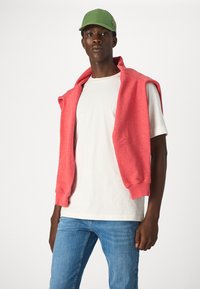 Czerwony bezrękawnik narzucony na biały t-shirt, zestawiony z jasnoniebieskimi dżinsami i zieloną czapką. Gładka tekstura i casualowy styl.