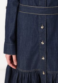 Robe en denim bleu foncé avec un ourlet en volants, dotée d'une fermeture à boutons et d'une taille ceinturée. Coutures visibles et léger éclat sur le tissu.