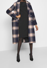 Rutig ullkappa i marinblått med orange och krämfärgade detaljer, med en dubbelsidig design och stora utknäppta kragar över en svart klänning.