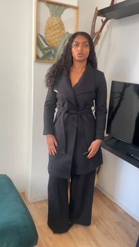 Mujer con cabello rizado y largo, vestida con un abrigo negro con cinturón y pantalones de pierna ancha, de pie en interiores cerca de una televisión y una obra de arte de piña.