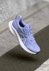 Αθλητικό παπούτσι σε ανοιχτό μωβ χρώμα με αντιανεμικό πλέγμα και υφή, λευκή σόλα και ανακλαστικές λεπτομέρειες, τοποθετημένο πάνω από μια επιφάνεια από τσιμέντο.