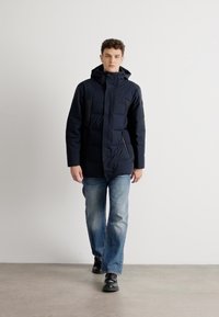 Giacca puffer blu navy con cappuccio, dotata di tasca con zip, patta frontale e design trapuntato. Indossata sopra jeans azzurri chiari e scarpe nere.