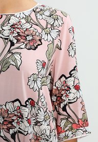 Light pink blouse featuring a floral pattern with white daisies and red accents. Short sleeves and a round neckline. Smooth texture.