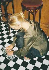 En person i en olivgrön blus med långa ärmar sitter på ett golv med svartvita plattor och hanterar en silverfärgad handväska med nitar.