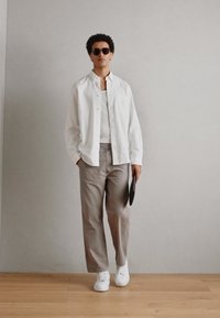Camicia bianca a bottoni, pantaloni grigi, sneakers bianche e occhiali da sole rotondi. Modello in piedi contro un muro chiaro con un pavimento in legno.