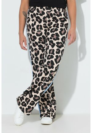 Osoba ubrana w beżowe spodnie z wysokim stanem z czarnym i brązowym wzorem w cętki oraz białe sneakersy, stojąca przy białej ścianie.