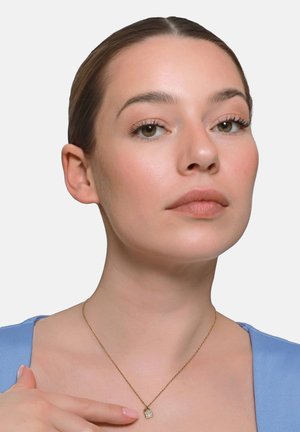 Frau mit zurückgebundenem Haar, die ein blaues Top und eine dünne goldene Kette mit einem quadratischen Anhänger trägt, vor einem einfarbigen grauen Hintergrund.