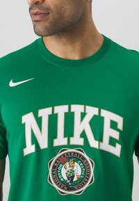 Grøn bomuld t-shirt med "NIKE" i hvid og guld skrift. Boston Celtics logo i midten. Rund hals, korte ærmer, glat tekstur.