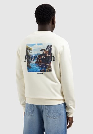 Mann trägt cremefarbenes Sweatshirt mit Küstenlandschaft und dem Text "Pure Path" auf dem Rücken, kombiniert mit hellblauen Jeans, blickt nach hinten.