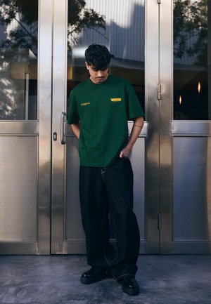 Mladi moški s temnimi kodrastimi lasmi, oblečen v zeleno majico in široke temne jeans hlače, stoji in gleda navzdol pred kovinskimi vrati stavbe.