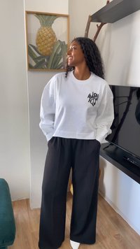 Sweatshirt curto branco com design gráfico preto, combinado com calças largas pretas. O ambiente inclui uma televisão e uma obra de arte com estampado de abacaxi.