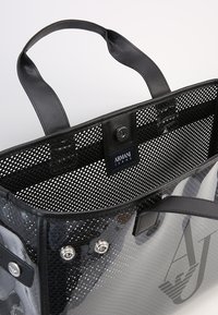 Průhledná černá síťovaná taška na rameno s hladkými černými koženými držadly a detaily. Má logo na nášivce a kovové patentky pro zapínání.