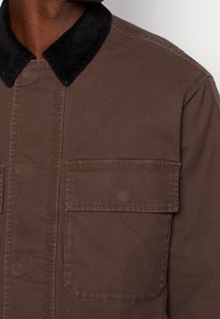 Brown jacket with large front pocket and black corduroy collar, worn by person with dark skin, visible from neck to chest.
