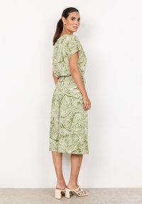 Kurzes grünes Kleid mit weißem Blattmuster, lockere Passform, kurze Ärmel, tailliert, knielange Länge, kombiniert mit cremefarbenen Blockabsatzsandalen.