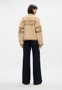 Beżowa, puszysta kurtka z kolorowymi wzorzystymi pasami na plecach i rękawach, połączona z ciemnymi, dzwonowymi dżinsami. Teksturowana tkanina, luźny krój.