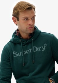 Turkusowy sweatshirt z kapturem, z sznurkami ściągającymi i kieszenią z przodu. Zawiera logo "SuperDry" w jaśniejszym odcieniu. Nałożony na koszulę w kratę.