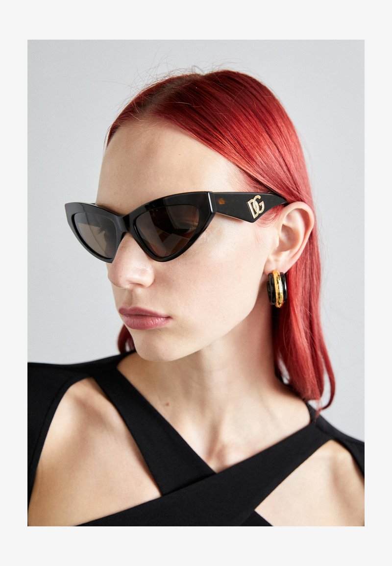 Mustad kassiirist kaitseprillid pruunide toonitud läätsedega ja kuldne logo tempeltes, koos suurte ümmarguste kuldsete ja mustade kõrvarõngastega.