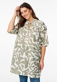 Tunika con motivo floreale in beige e oliva, con scollatura a chiave, dettaglio arricciato sulle spalle e maniche a palloncino corte. Realizzata in tessuto morbido.