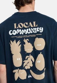 Κότινη μπλε βαμβακερή μπλούζα με μπεζ εικονογραφήσεις φρούτων που έχουν εκφραστικά μάτια και κείμενο που γράφει "ΤΟΠΙΚΗ ΚΟΙΝΟΤΗΤΑ" σε διάφορες γραμματοσειρές.