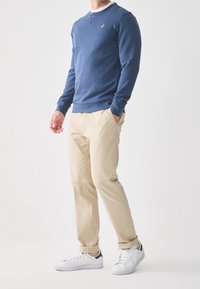 Mandlig model iført en blå langærmet sweatshirt, beige chinos og hvide sneakers, stående mod en ensfarvet hvid baggrund.