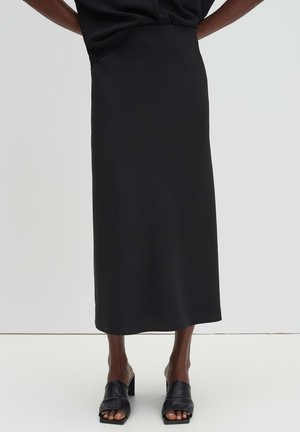 Jupe noire longueur cheville portée avec des mules à talons ouvertes noires, debout sur un fond blanc uni.